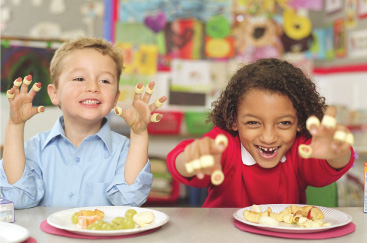 Two smiling elementary school students sit at a table with lunch on their plates; they show off rings of pasta around each of their fingers.