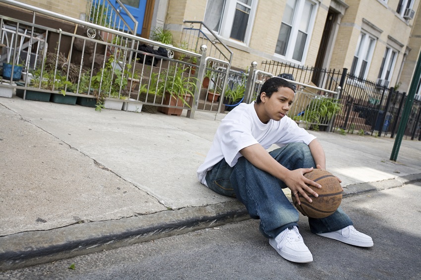 Young boy holds a basketball and sits on the sidewalk.
