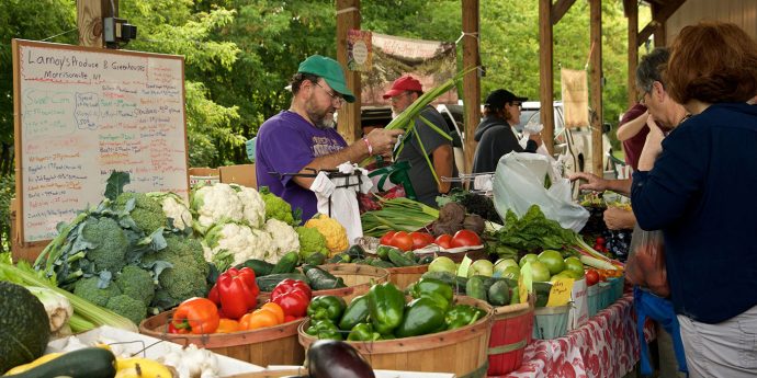 A fruit and vegetable stand at a farmers market with vendors and shoppers.