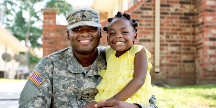 A male veteran of dark skin tone wearing military fatigues smiles with his daughter.
