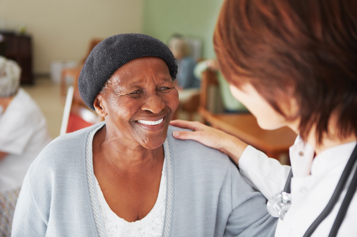 A female health care provider puts a comforting hand on an elderly female patient who is smiling.