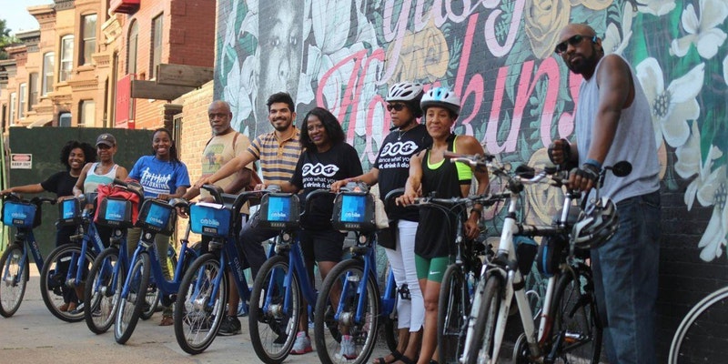 A group of New Yorkers with medium to dark skin tone stands with CitiBikes in front of a mural of art.