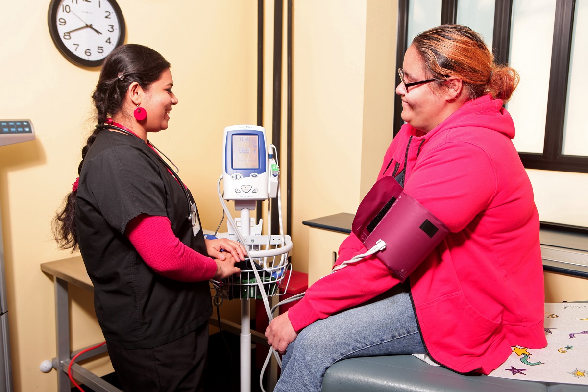A female health care provider with medium-dark skin tone takes the blood pressure of a female patient with medium skin tone.