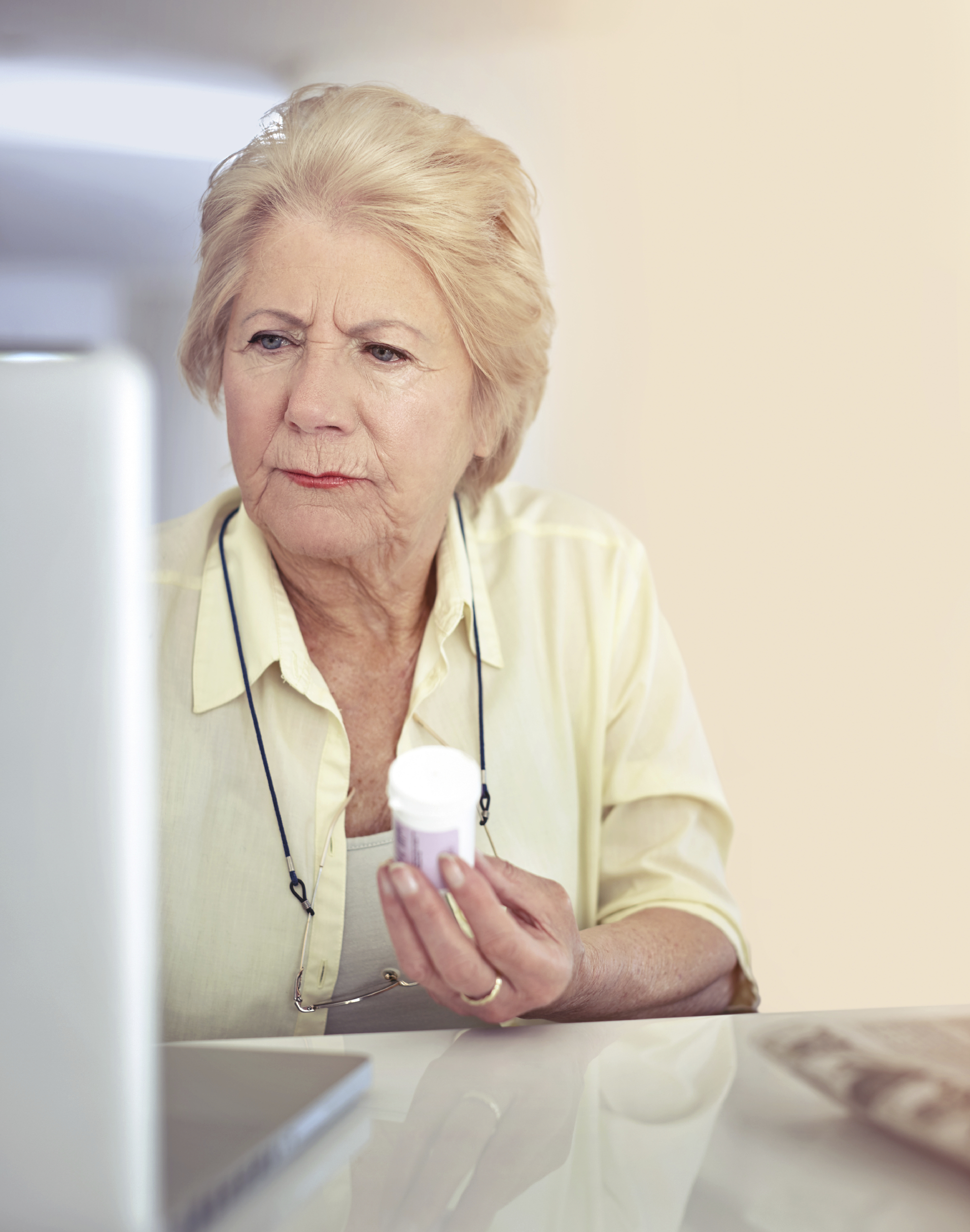 Elderly woman holds a prescription bottle and looks at a computer.