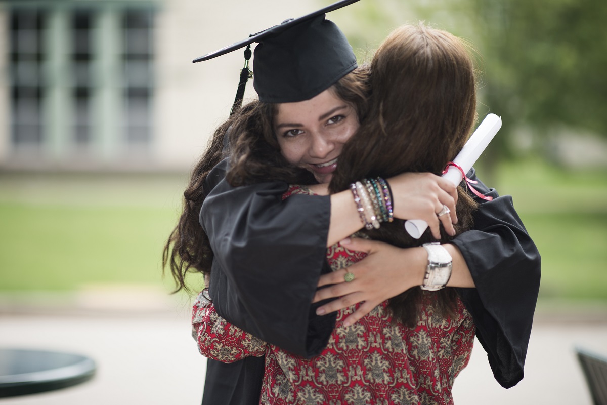 A student of medium-light skin tone wears a graduation cap and gown, hugging a family member.