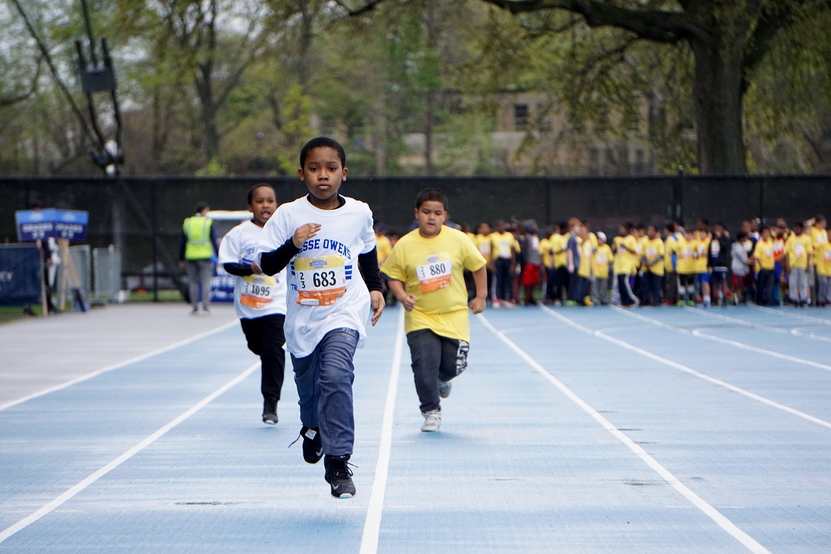 Three children are running a race at a track, with a throng of other kids lined up behind them.