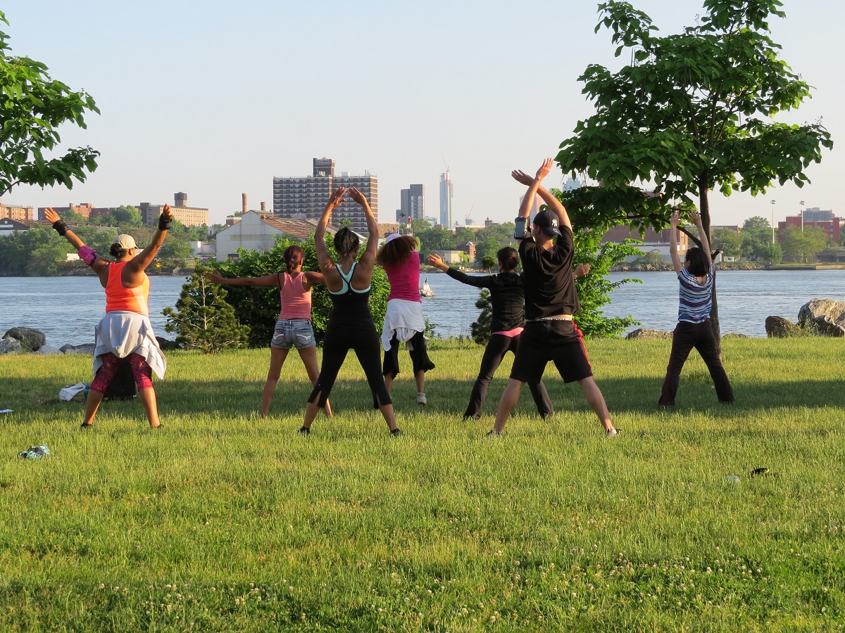 Participants in an outdoor exercise class stretch in a riverfront park.