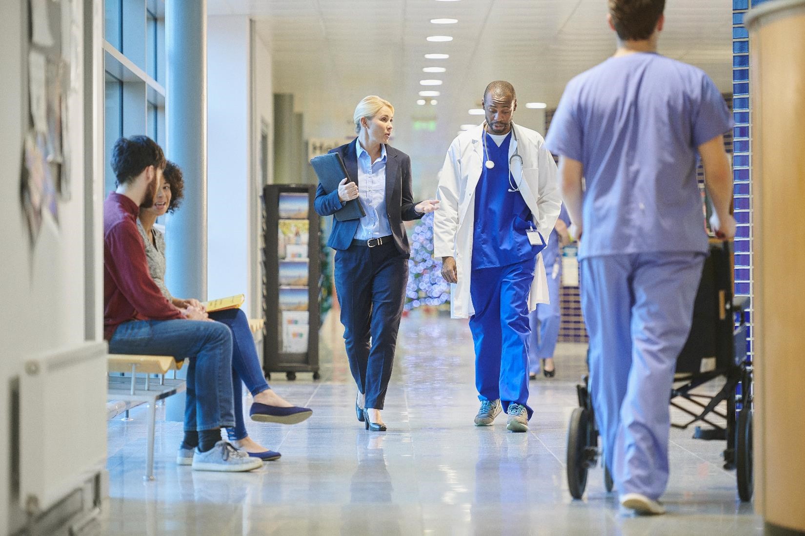 A doctor wearing scrubs and a white coat walks down a hospital hallway chatting with a hospital administrator while other staff, patients, and visitors are visible in the background.
