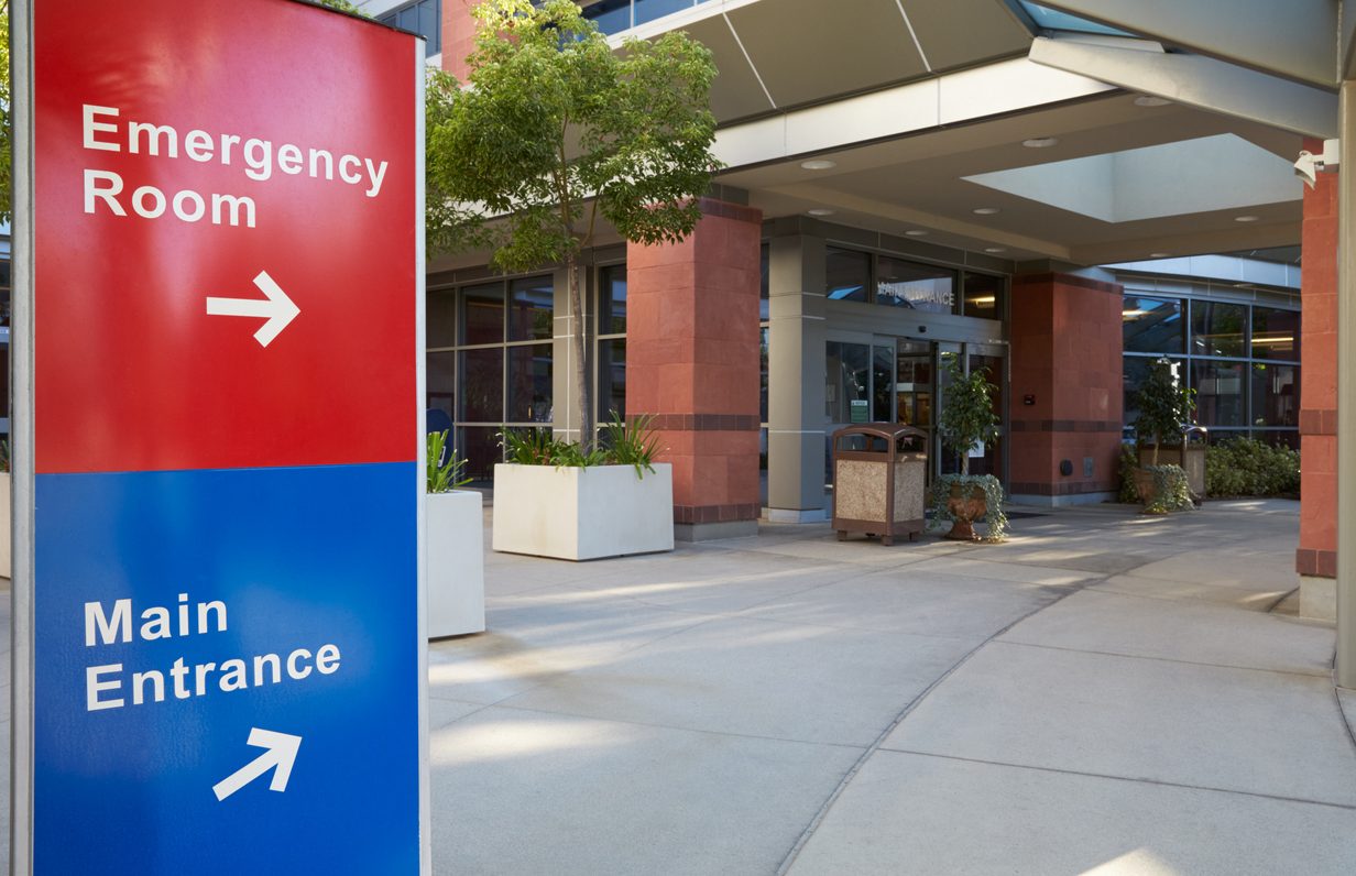 The front of a modern hospital building with directional signs pointing to the emergency room and the main entrance.