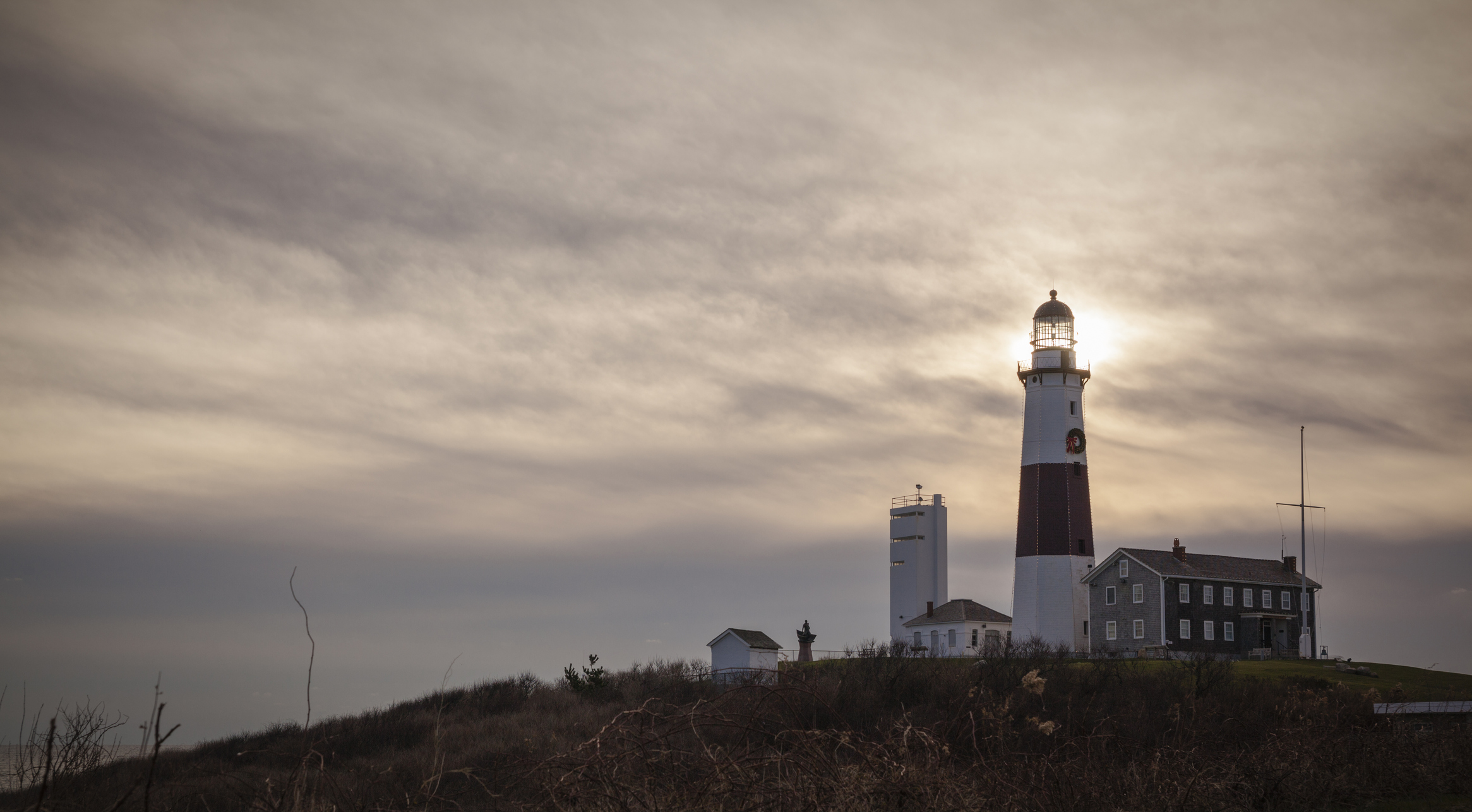 A lighthouse at the top of a hill on a gloomy day, sun barely poking through a sky of gray clouds.