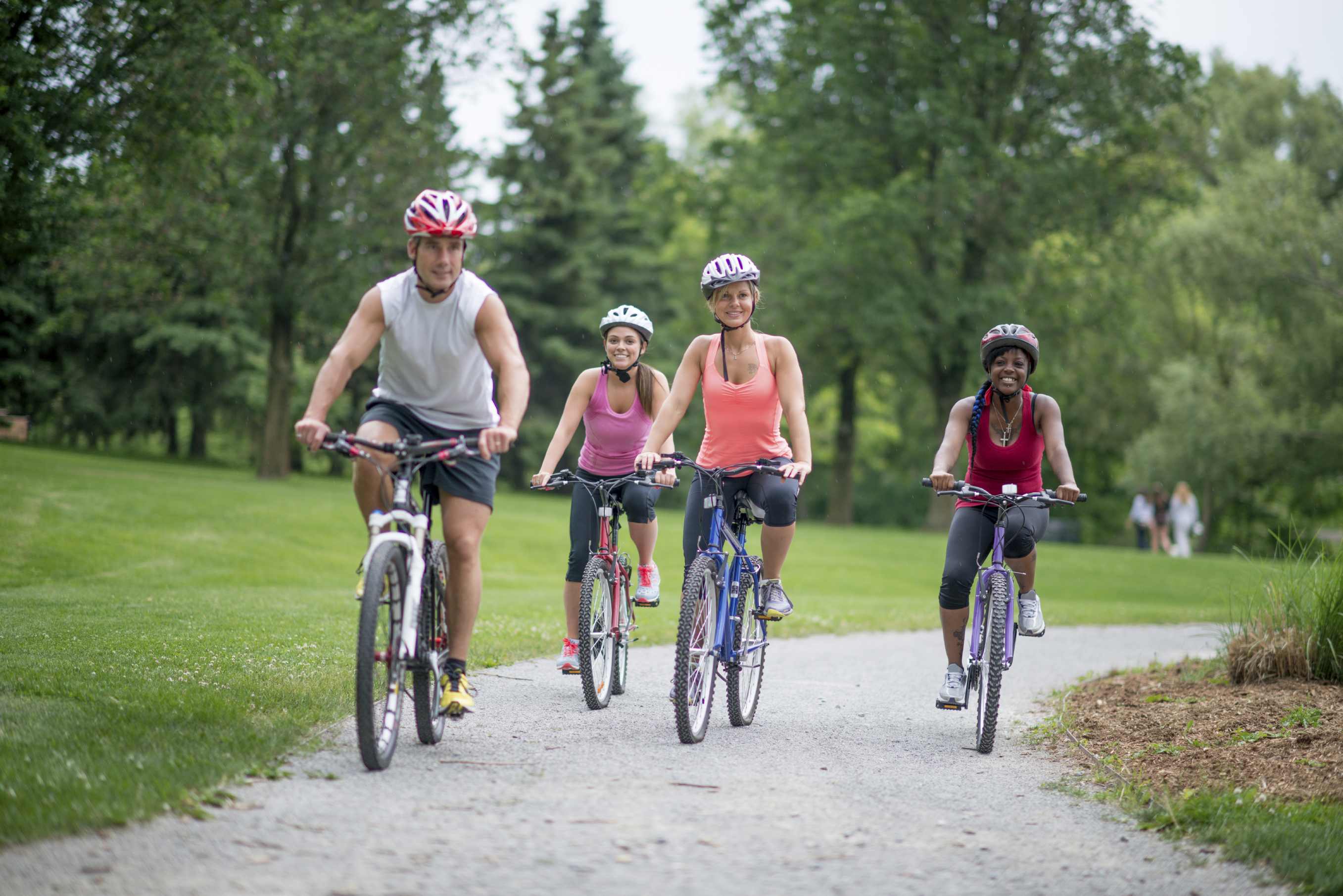 Four smiling people wearing helmets as they bicycle on a path through a park.