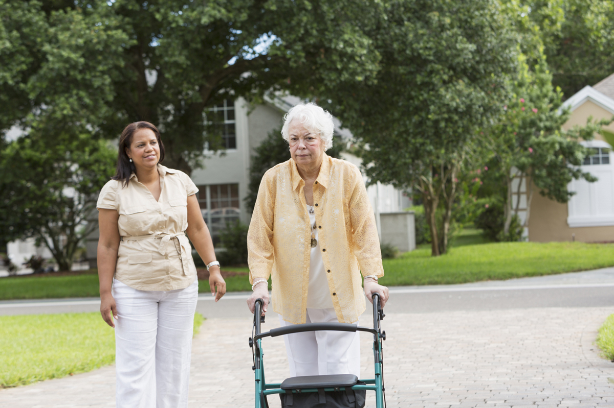 An older person using a walker takes a walk around her neighborhood, accompanied by a caregiver.