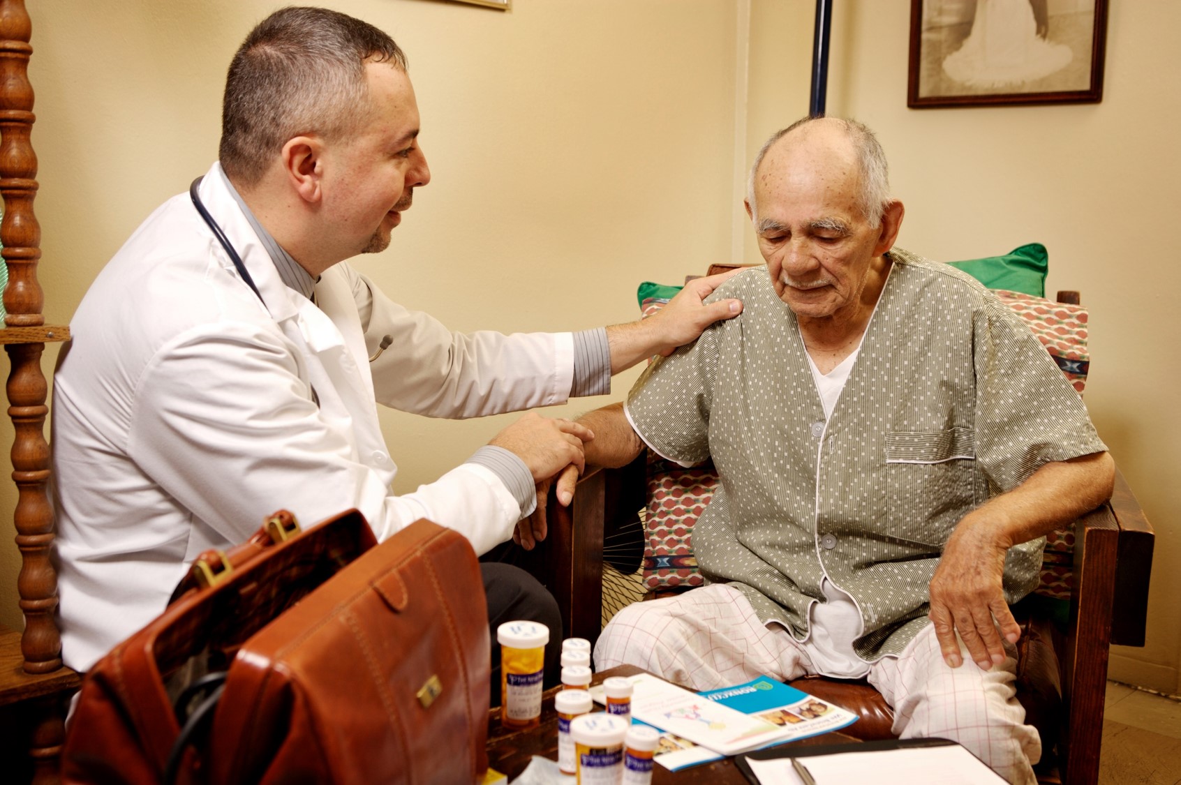 A doctor in a white coat sits next to an elderly patient in his living room, placing one hand kindly on his shoulder and the other on the patient's hand. Prescription bottles are scattered on the coffee table in front of them.