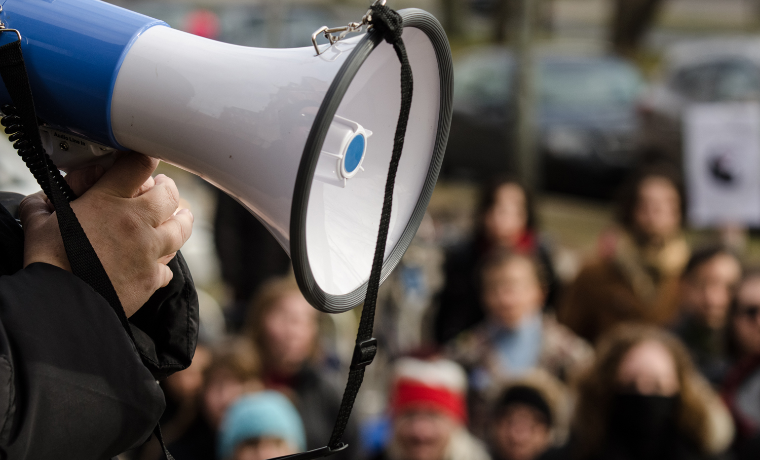 A hand holding a megaphone in front of a blurred background of people at a rally or protest.