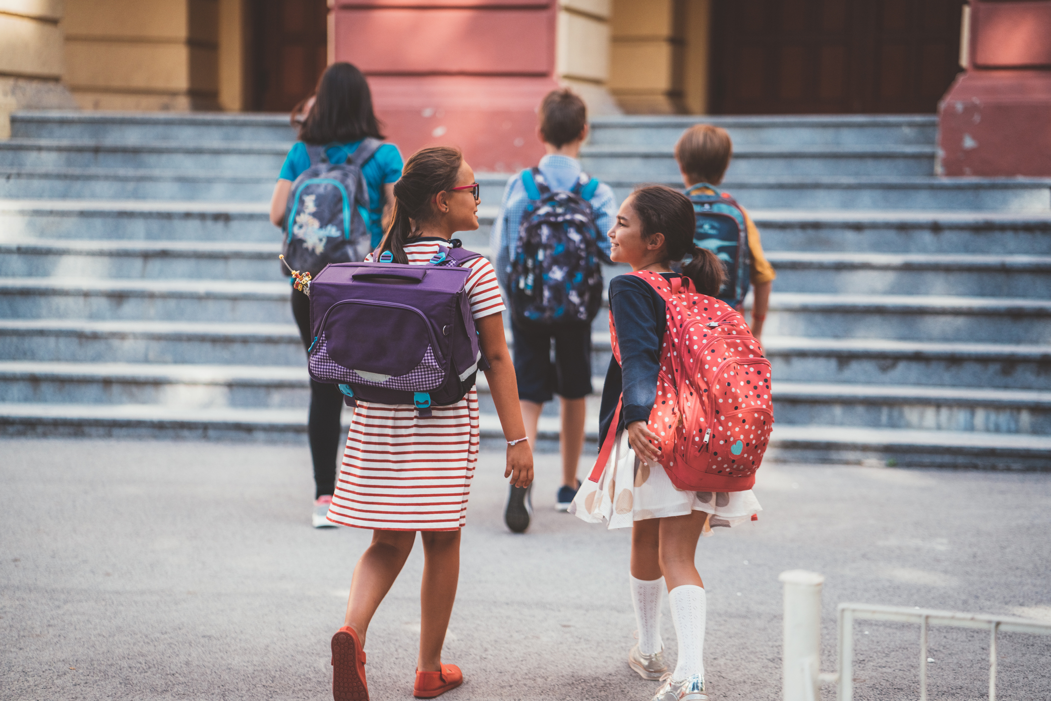 Two adolescent girls wearing backpacks walk toward a middle school building with other students in backpacks.