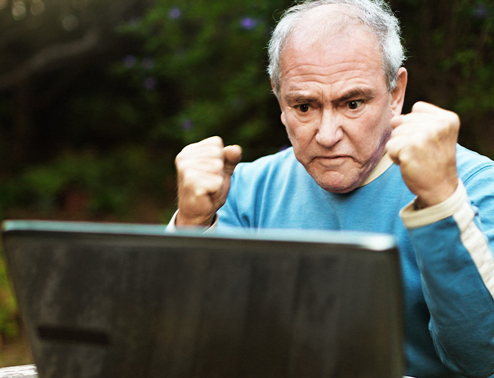 An elderly man holds up his fists in frustration in front of a computer.
