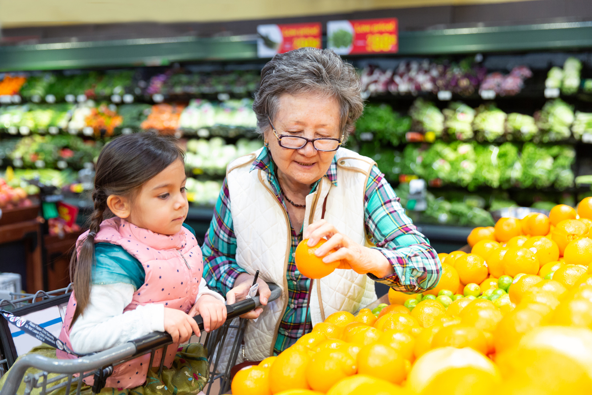 A grandmother with medium skin tone grocery shops with her granddaughter of medium skin tone.