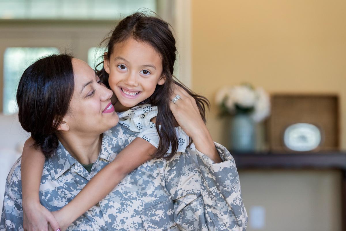 A female veteran of medium skin tone smiles at her young daughter behind her.