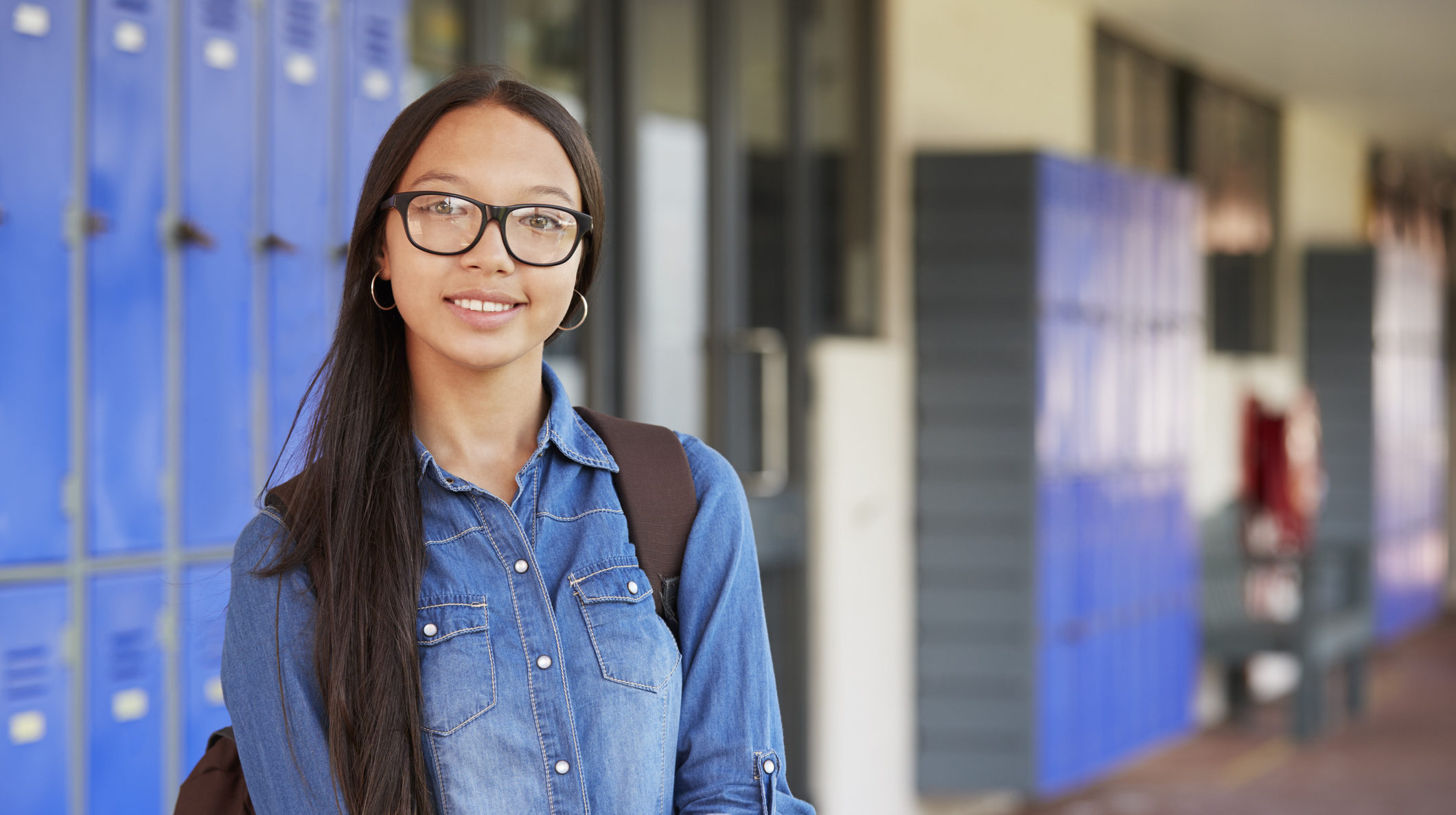 An adolescent girl wearing glasses and a backpack smiles in front of a set of lockers at school.