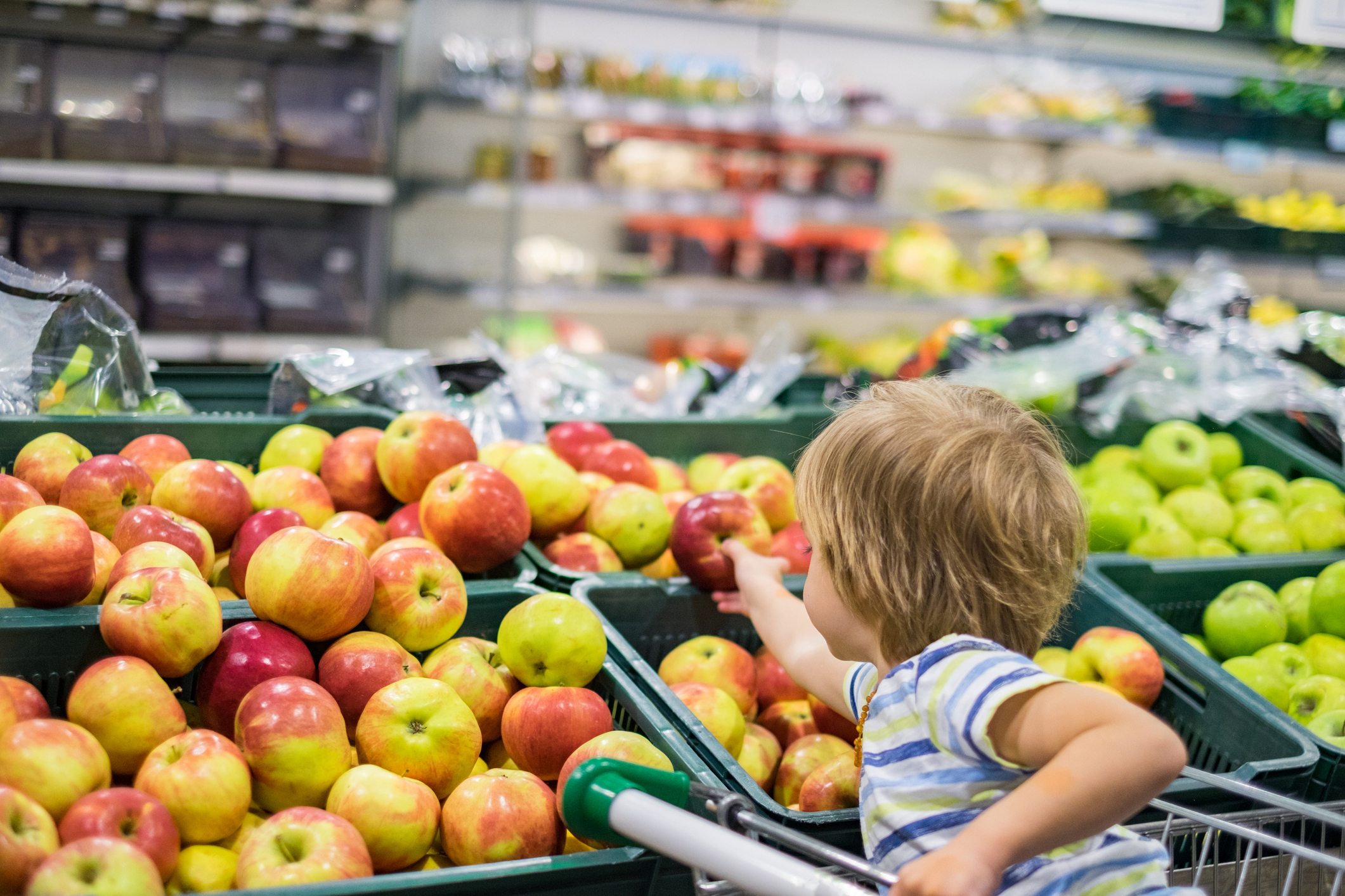 A young child with light skin tone leans over from his seat in a grocery cart to pick an apple.