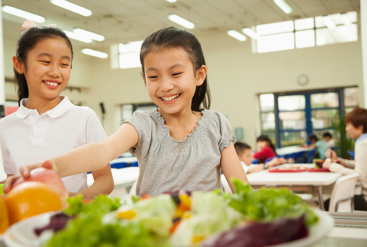Two smiling students with medium skin tone reaching for fruit in a school cafeteria.