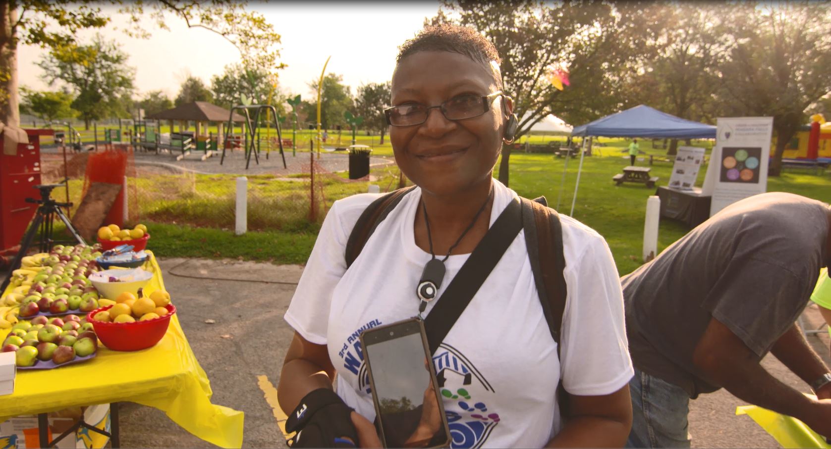 A smiling woman with dark skin tone poses in front of a fruit stand at the Walk Niagara Falls annual event.