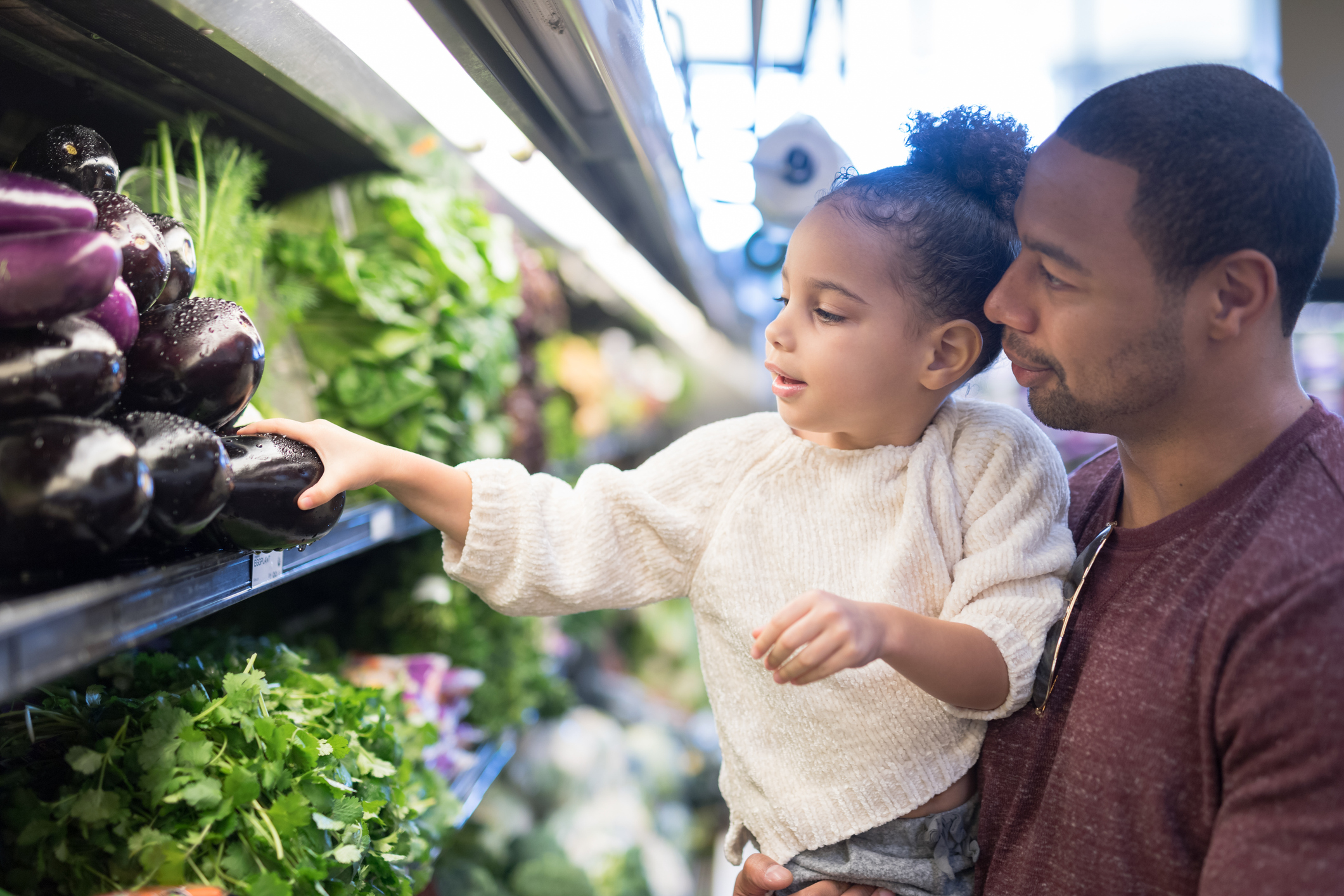 A father with medium-dark skin tone hold his daughter with medium skin tone at a grocery store while she picks out produce.