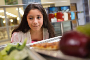 A middle school student pressing her lips together in a small smile as she crouches down and leans in to look at the cafeteria lunch options.