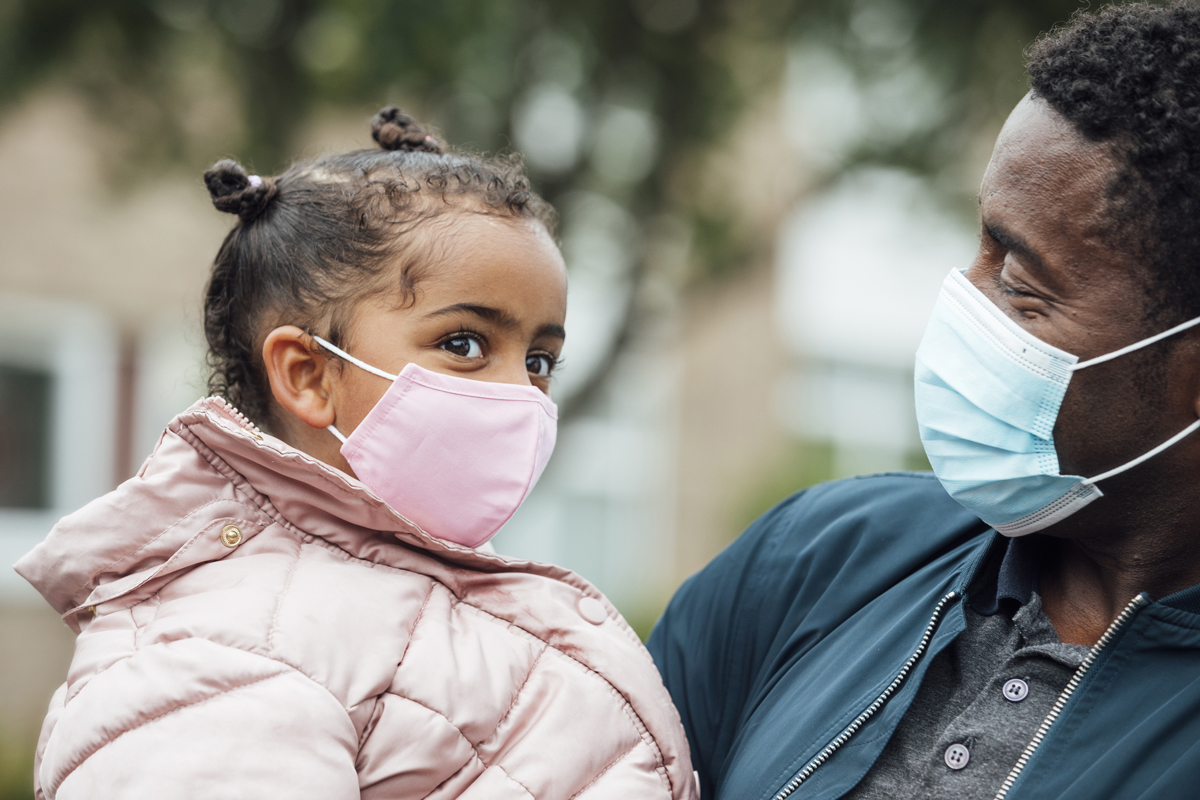 A father wearing a protective mask smiles and holds his young daughter who is also wearing a protective mask.