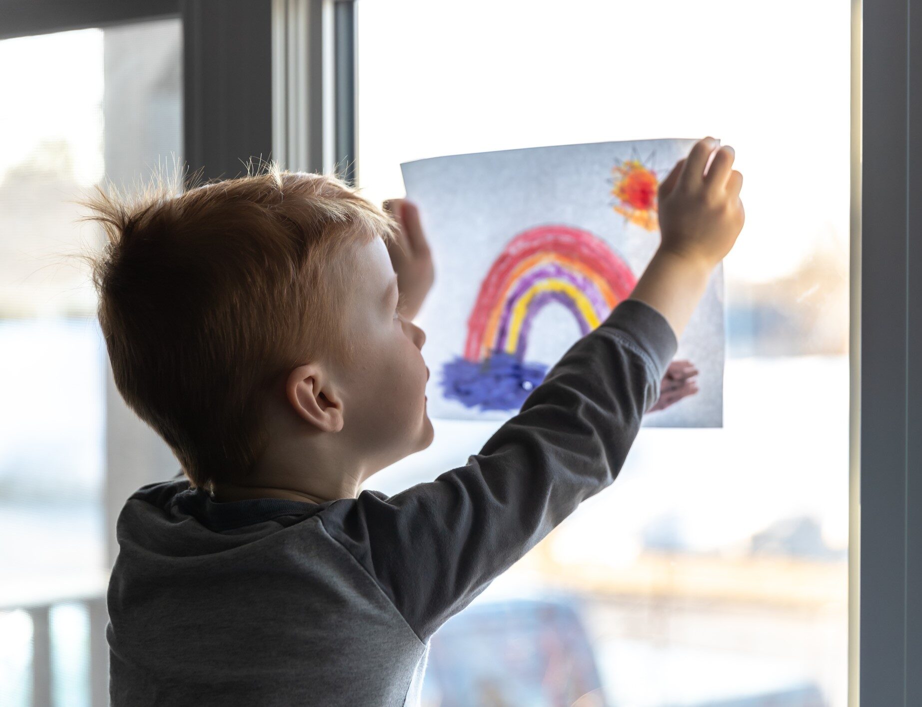 A young boy holds up a drawing of a rainbow against a window.