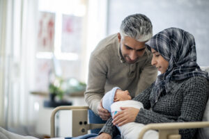 A mother with medium skin tone wearing a head scarf and a father with medium skin tone hold their newborn baby in a hospital.