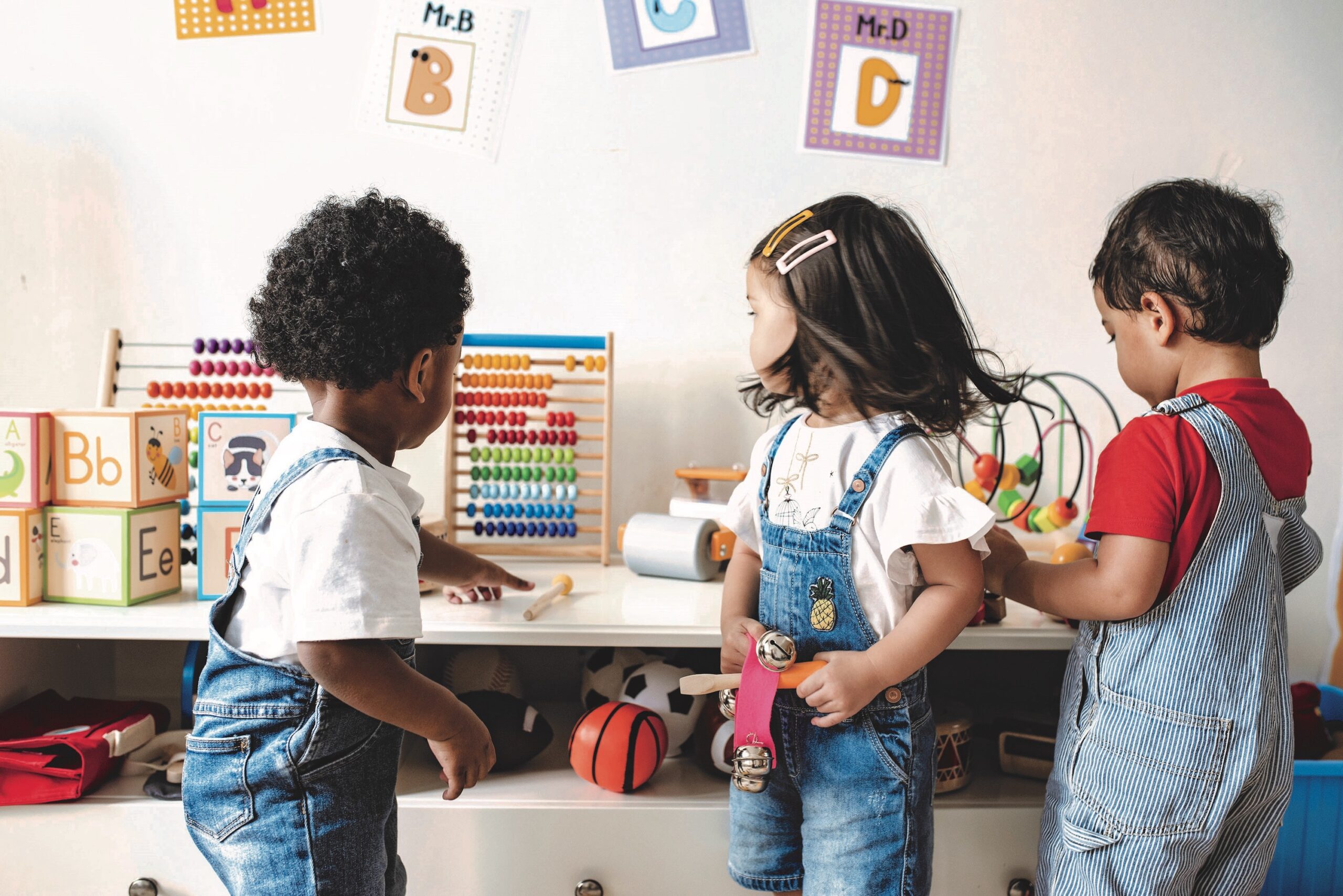 Three young children of light, medium, and dark skin tones play with toys at school.
