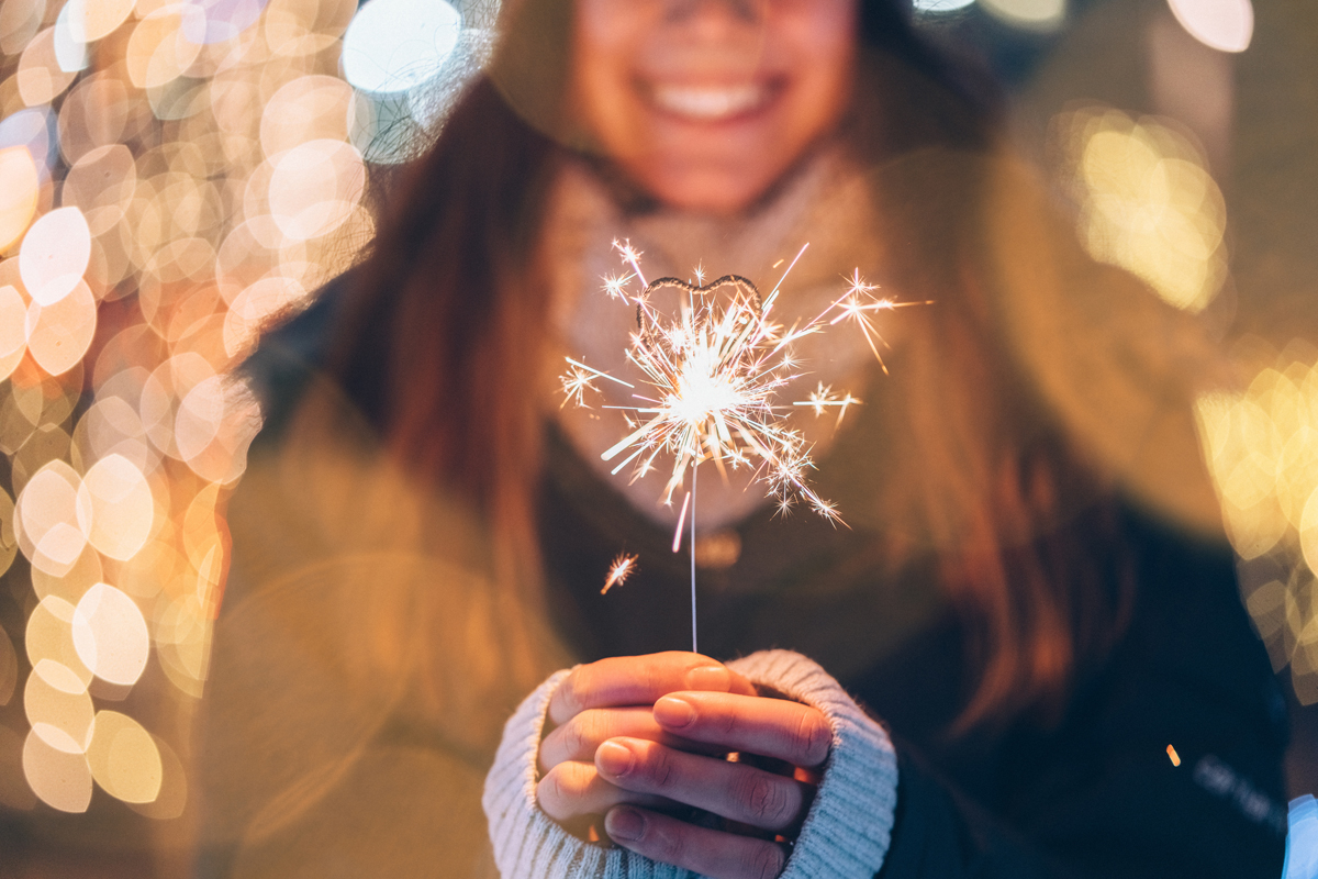 A smiling person holding out a lit sparkler in front her of.