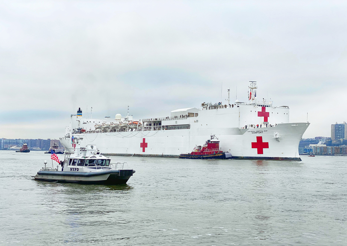 A large white U.S. Navy hospital ship emblazoned with three red crosses sails into New York City. A much smaller New York Police Department boat is to its left, with an American flag sailing from it.