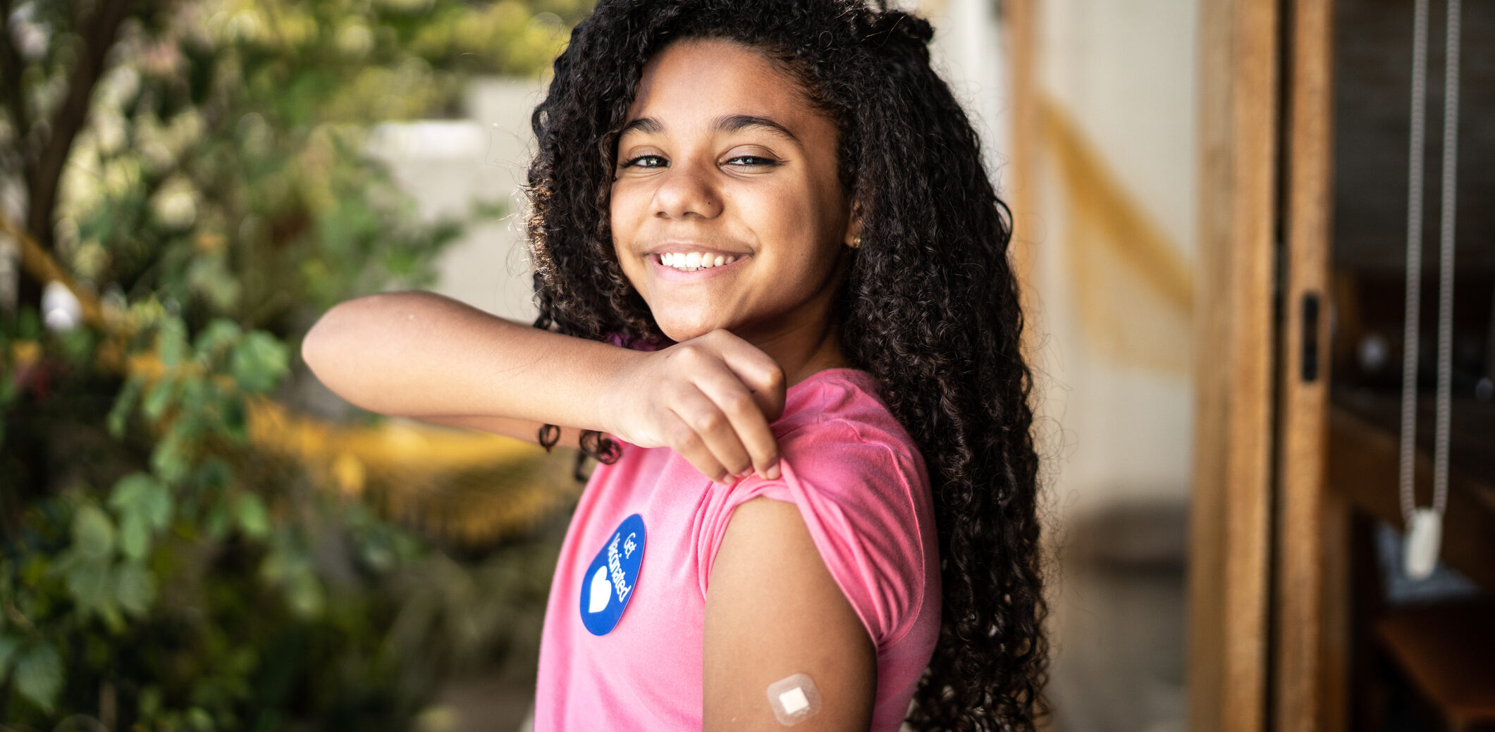 A smiling adolescent with medium skin tone rolls up the sleeve of a pink t-shirt to show off a small band-aid on the left arm. A sticker on the t-shirt reads "Get Vaccinated."