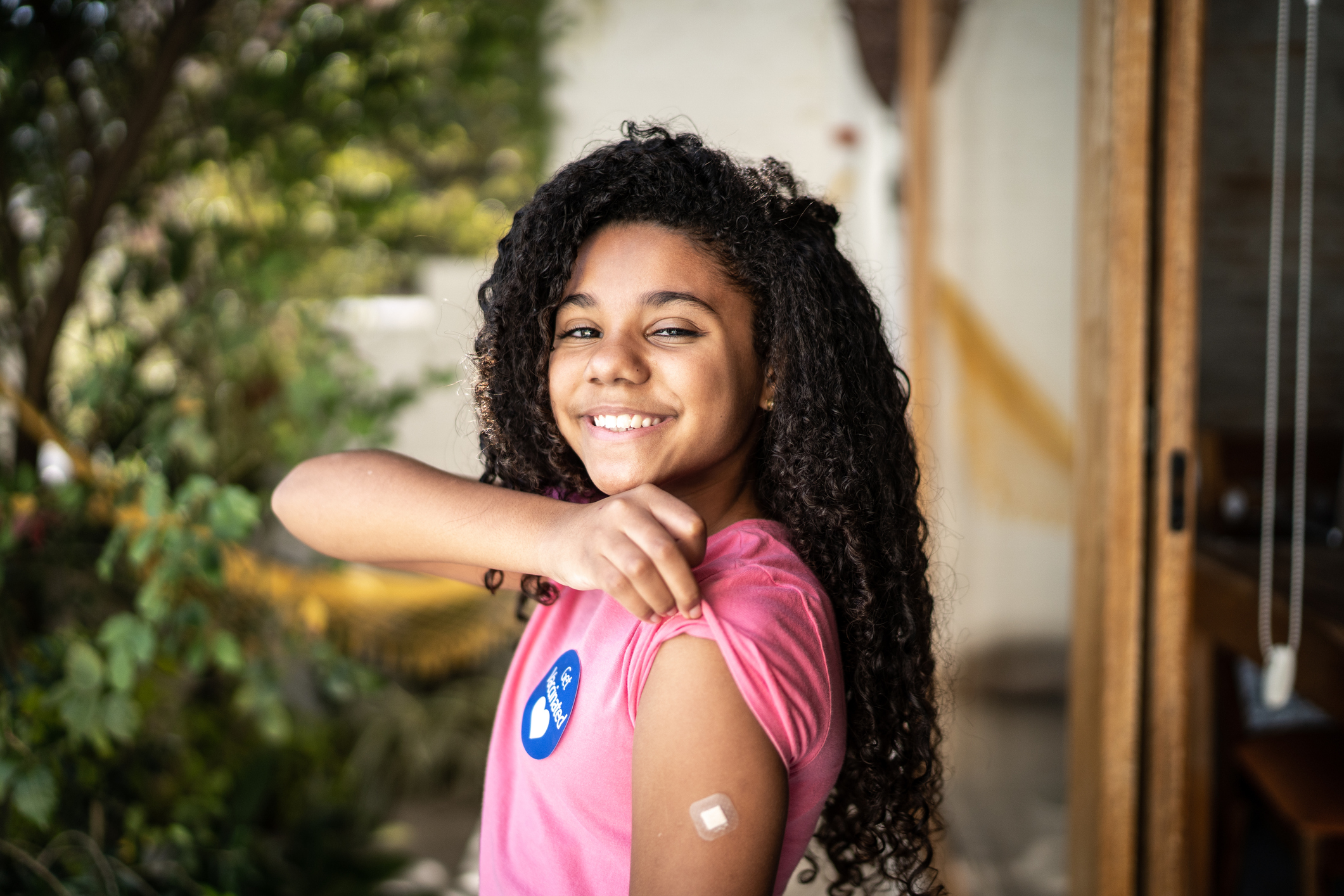 A smiling adolescent with medium skin tone rolls up the sleeve of a pink t-shirt to show off a small band-aid on the left arm. A sticker on the t-shirt reads "Get Vaccinated."