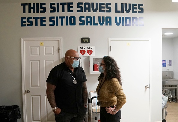 Director of overdose prevention center talking to worker. Behind them, painted letters on the wall spell out "This site saves lives" and, beneath that, "Este sitio salva vidas."
