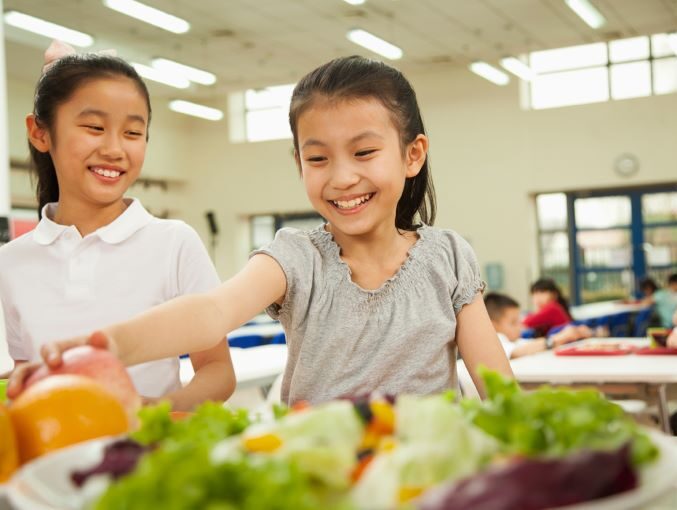 Two smiling students with medium skin tone reaching for fruit in a school cafeteria.