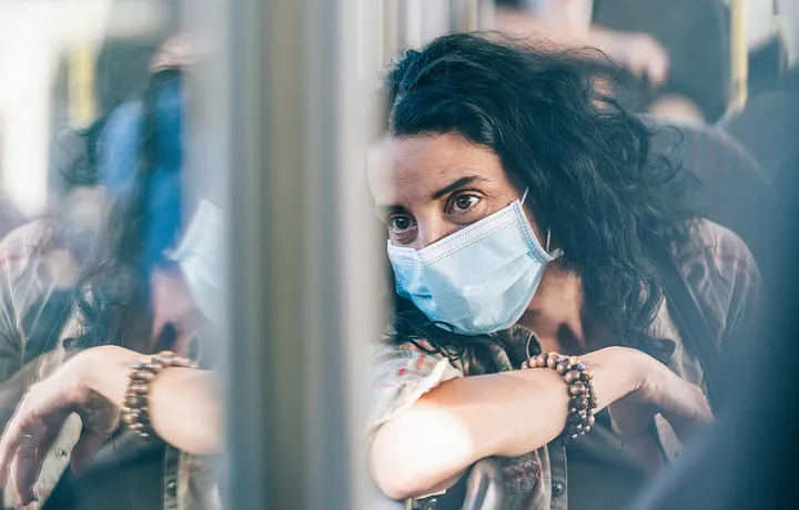 A woman wearing a surgical face mask rides public transportation and looks out of the window.