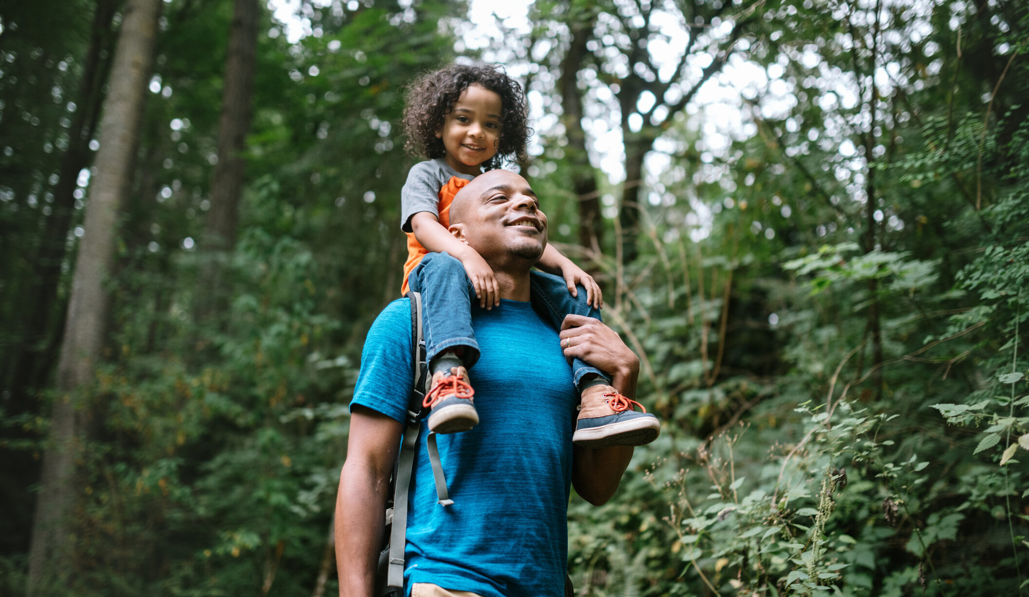 A father with medium-dark skin tones carrying his son on his shoulders through a forest.